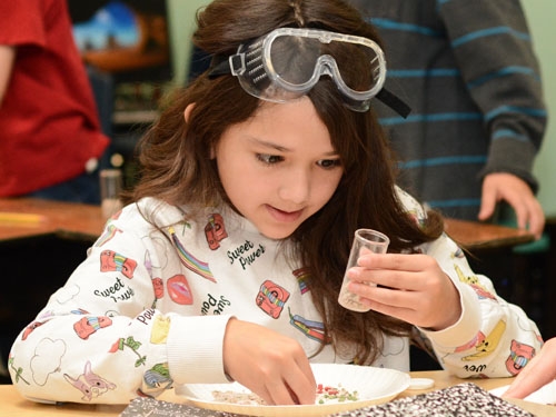 young girl with long brown hair, white sweatshirt, and safety goggles on her forehead looking at a vial in her left hand as she puts rocks inside of it