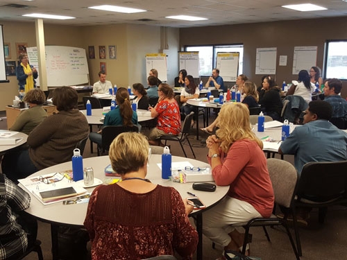 Science teachers sitting at round tables in large classroom attentively listening to institute leader.