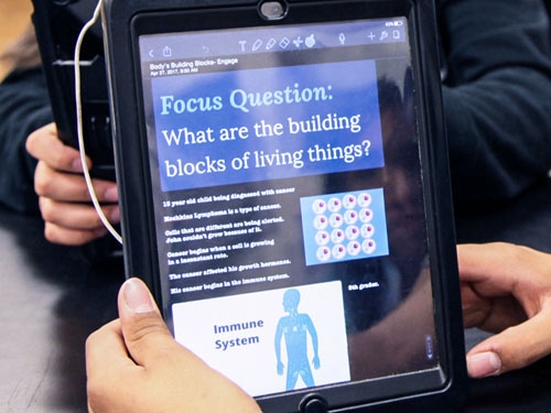 Student holding an iPad with a lesson on it called "What are the building blocks of living things?"