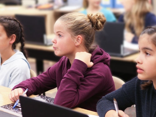 Girl with blonde hair tied in a bun and burgundy hoodie sitting at a table in classroom with other girls.